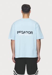 Světle modré bavlněné tričko s černým a bílým textem "PEGADOR Next Wave" na zádech; volný střih a krátké rukávy.