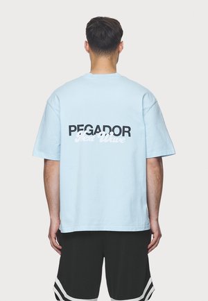 Světle modré bavlněné tričko s černým a bílým textem "PEGADOR Next Wave" na zádech; volný střih a krátké rukávy.