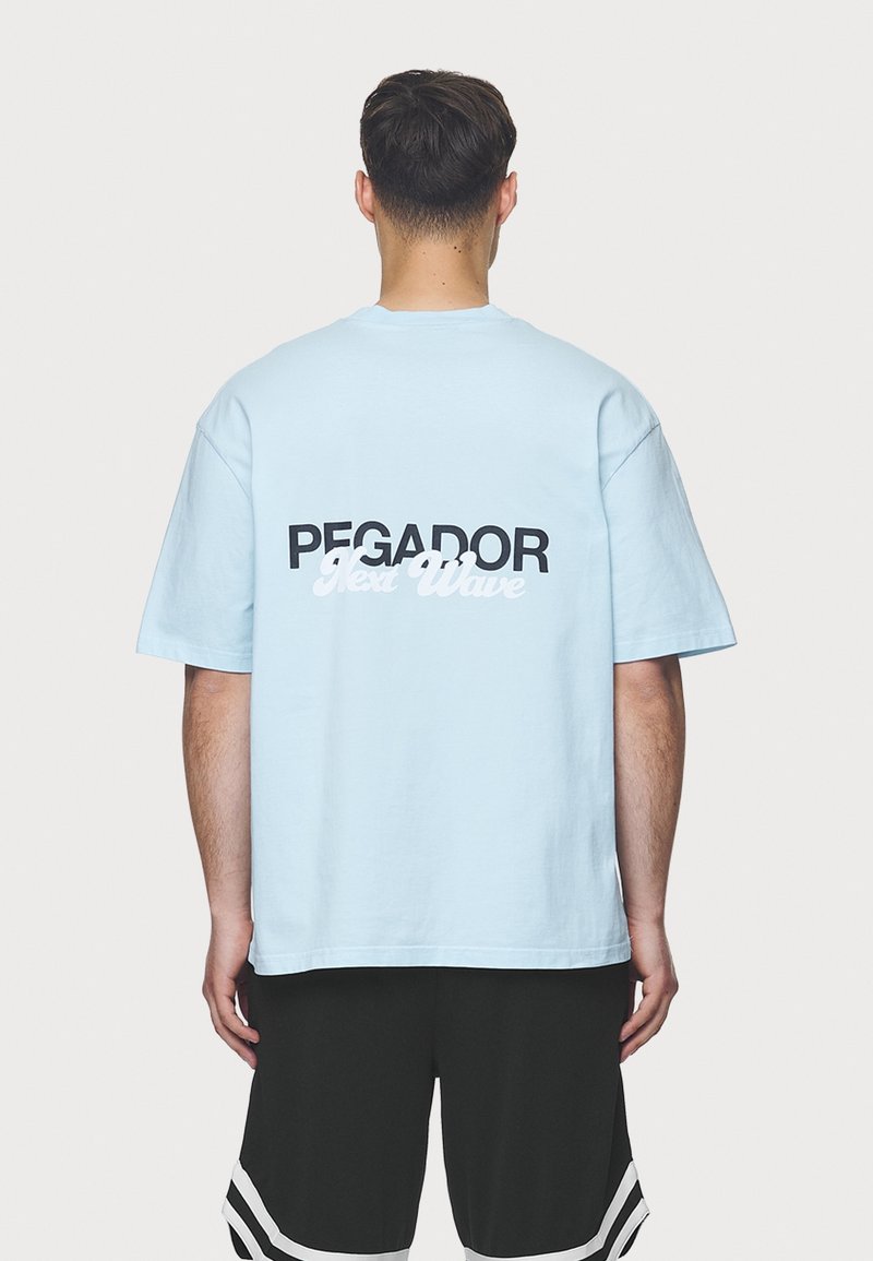 Světle modré bavlněné tričko s černým a bílým textem "PEGADOR Next Wave" na zádech; volný střih a krátké rukávy.