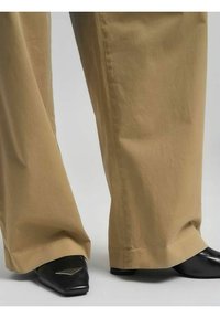 Pantaloni beige a gamba larga con una consistenza liscia e senza pieghe, abbinati a scarpe nere con punta appuntita e un accento metallico sulla punta.