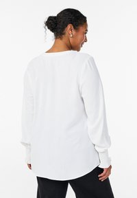 Mujer con el cabello rizado y oscuro recogido en un moño, que lleva pendientes plateados y una blusa blanca de manga larga con puños fruncidos, vista de perfil sobre un fondo blanco.