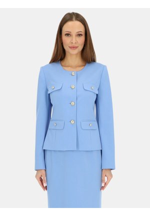 Kobieta ubrana w dopasowany jasnoniebieski garnitur z złotymi guzikami i kieszeniami z patkami, stojąca na tle jednolitego jasnego tła.