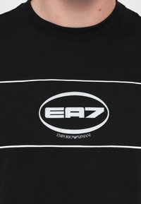 Černé tričko s krátkým rukávem s bílými vodorovnými pruhy. Uprostřed logo s nápisem "EA7" uvnitř oválu a text "Emporio Armani" pod ním.