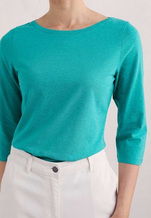 Femme portant un haut bleu sarcelle à manches trois-quarts rentré dans un pantalon blanc taille haute avec une fermeture à boutons devant.