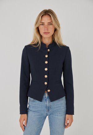 Donna bionda che indossa un blazer blu navy aderente con bottoni dorati e jeans azzurri, in piedi davanti a uno sfondo grigio chiaro uniforme.