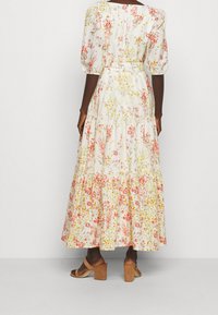 Kvinna i en vit blommig maxiklänning med puffärmar och beige klacksandaler med blockhälar, som står mot en enkel ljus bakgrund.