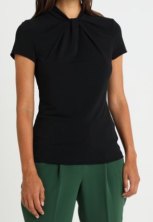 Haut noir à manches courtes avec un détail de col plié, texture lisse, forme ajustée, associé à un pantalon large vert.