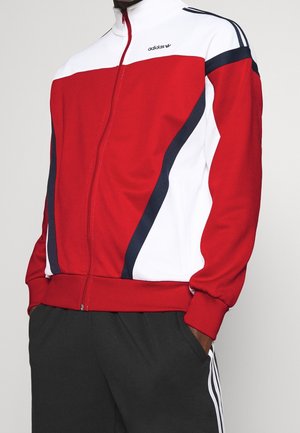 Mann trägt eine rot-weiß-schwarze Adidas-Zip-up-Jacke mit schwarzen Hosen, die weiße Seitenstreifen haben, Hände in den Taschen.