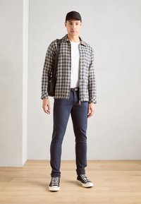 Camisa de xadrez em azul e amarelo, t-shirt branca, calças azul-marinho, sapatilhas pretas e boné preto; modelo segurando uma bolsa de ombro preta.
