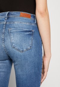 Jeans de mezclilla azul ajustados que presentan una cintura alta, una etiqueta de marca de cuero marrón y bolsillos traseros con una sutil textura desgastada.