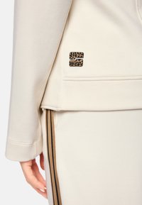 Cremefarbener Sport-Hoodie mit einem braunen Leopardendruck-Logo. Ausgestattet mit einer glatten Textur und gerippten Bündchen, kombiniert mit Jogginghosen mit braunen und schwarzen Streifen.