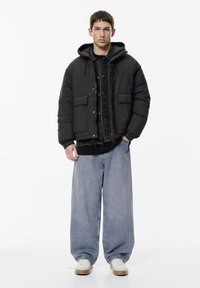 Jachetă puffer neagră cu glugă și buzunare frontale, purtată peste o cămașă neagră cu butoni, completată cu blugi evazați de culoare albastră deschisă și adidași albi.