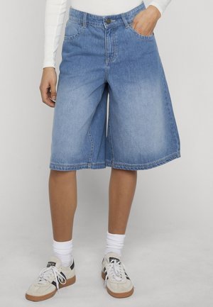 Inimene, kes kannab keskpikkuses tuhmiks kulunud siniseid teksapükse taskutega, valgeid sokke, valgeid tossusid ja valget pikkade varrukatega tuba.