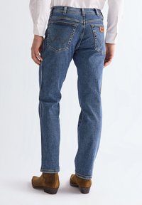 Kék farmert és barna velúr bokacsizmát viselő férfi hátulnézetből, egyszínű fehér háttér előtt.