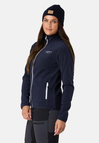 Tummansininen fleece-takki korkealla kauluksella, jossa on kaksi valkoista vetoketjullista taskua ja logo rinnassa; yhdistettynä tummiin housuihin ja tummansiniseen pipoon.