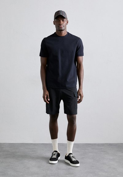 Μαύρο κοντομάνικο υφασμάτινο μπλουζάκι, μαύρο cargo σορτς, λευκές ριγές κάλτσες, μαύρα αθλητικά παπούτσια με λευκή σόλα, και καπέλο με πολύχρωμες ρίγες.