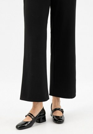 Des chaussures noires en cuir verni avec un bout carré, deux brides à la cheville et un talon carré, portées avec un pantalon noir fluide.