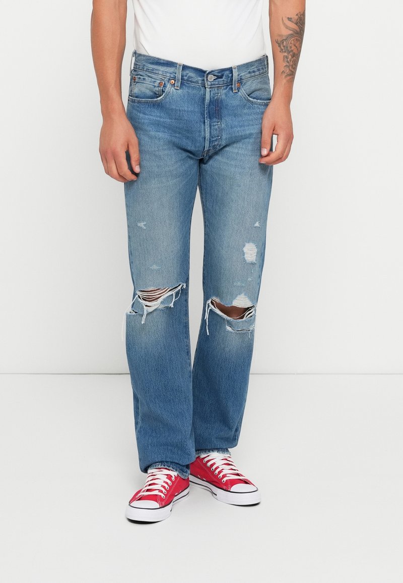 Man som bär ljusblå söndriga jeans, röda sneakers och en vit skjorta, stående mot en enfärgad vit bakgrund.