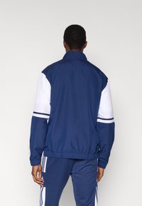 Giacca con zip blu navy con maniche bianche. Vita e polsini elasticizzati, con strisce a contrasto sulle maniche. Materiale resistente e leggero.