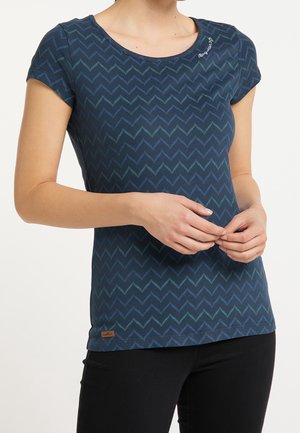 Mujer con camisa azul marino de manga corta con patrón de zigzag en color verde azulado, pantalones negros, manos relajadas entrelazadas a la altura de la cintura.