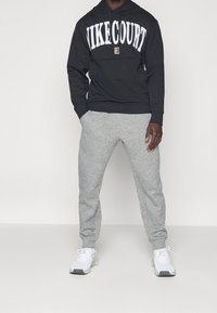 Sudadera con capucha negra con el logo "NIKE COURT", combinada con pantalones deportivos gris claro y zapatillas atléticas blancas. Diseño sencillo y relajado.