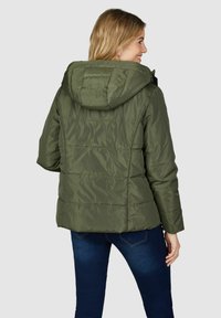 Veste matelassée vert olive avec une capuche doublée, arborant une texture piquée et un design de dos emmanché, accompagnée d'un jean bleu foncé.