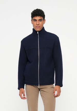 Navy ull zip-jacka med hög krage, som har en slät textur och strukturerade axlar. Silverfärgad dragkedja som sträcker sig nerför framsidan.