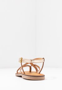 Sandalo in pelle in tono naturale con cinturini incrociati, fibbia alla caviglia, suola marrone e dettagli intrecciati sulla parte superiore. Design semplice, stile piatto.