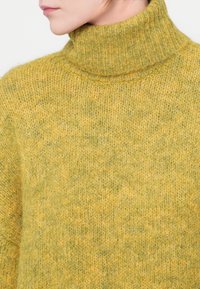 Hořčicově žlutý pletený rolák s jemnou texturou, který má ribbed akcenty a uvolněný střih. Je doplněn o jemný zelený nádech.