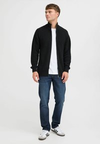 Svart ribbet hettejakke med glidelås, tettsittende design, brukt over en hvit t-skjorte, kombinert med blå jeans og grå/hvite joggesko.