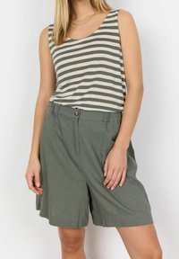 Žena v bezramínkovém zeleno-bílém pruhovaném topu zastrčeném do volných olivově zelených šortek na bílém pozadí.