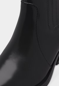 Κοντινή λήψη μαύρου λουστρινένιου δερμάτινου μποτάκι αστραγάλου με ελαστική πλευρική επιφάνεια και ορατές ραφές.