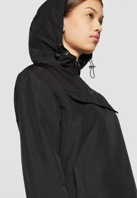 Svart vattentät jacka med huva, dragkedja fram och elastiska detaljer. Har en ficka med klaff och en stilren design med slät, glänsande yta.