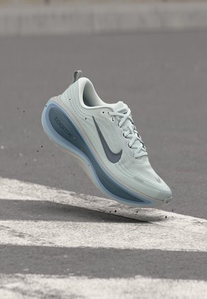 Chaussure de course Nike gris clair avec semelle bleue en l'air au-dessus de l'asphalte et des marquages routiers blancs, montrant un mouvement avec de petits débris en dessous.
