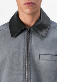 Κοντινό πλάνο ενός ατόμου που φοράει γκρι μπουφάν με μαύρο δερμάτινο γιακά και κεντρικό φερμουάρ, δείχνοντας ένα μέρος του στήθους και του στόματος.