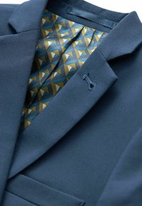 Blazer blu navy con rever a punta; l'interno presenta un motivo geometrico in blu, oro e crema. Dettaglio ricamato sul rever.