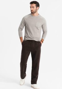 Harmaa pitkähihaisen paita etutaskulla, yhdistettynä tummanruskeisiin housuihin. Mallilla on valkoiset lenkkarit, mikä tuo esiin rentoa tyyliä ja yksinkertaista muotoilua.