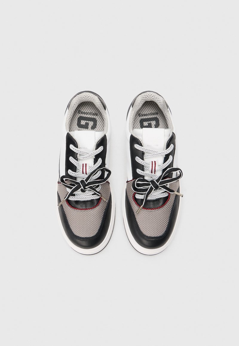 Baskets de sport noires et grises en matériaux mesh et cuir, à bout rond, semelle blanche plate et lacets noirs. Couture accent rouge visible.