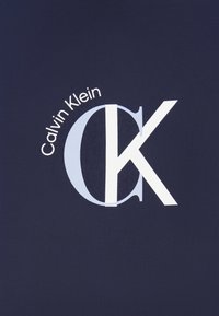 Tumši zils audums ar lielu baltu "CK" logotipu un virs tā baltā krāsā apliektu tekstu "Calvin Klein". Gluda tekstūra, moderns dizains.
