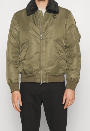 Homme portant une veste bomber vert olive avec un col en fausse fourrure noire et un pantalon noir, debout devant un fond uni.