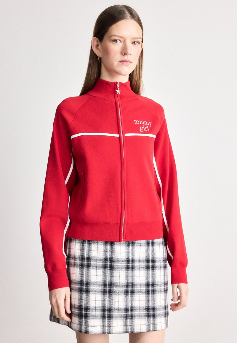 Chaqueta roja con cremallera y detalles en blanco, con la marca "tommy girl". Cuenta con un cuello alto y mangas largas, combinada con una falda de cuadros en blanco y negro.