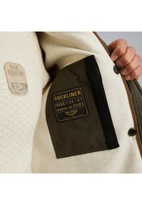 Unutrašnjost jakne u sivo-smeškanom i maslinasto zelenom tonu s teksturiranom kremastom podlogom. Ima crni džep i prišivene oznake s informacijama o proizvodu.