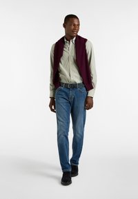 Un hombre lleva una camisa a rayas verde y blanca, con un chaleco de punto morado oscuro, jeans azules y zapatos oscuros.