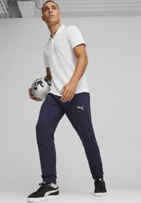 Witte polo, marineblauwe trainingsbroek, houdt een zwart-wit voetbal vast. Bevat Puma-logo op de broek. Sneakers met zwart-witte accenten.