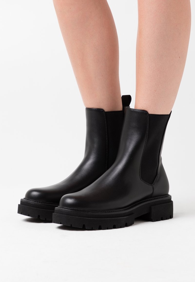 Bottes chevilles en cuir noir avec semelles épaisses portées sur jambes nues sur un fond blanc.