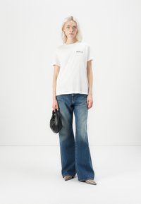 Witte katoenen t-shirt met de tekst "REPLAY", gecombineerd met lichtblauwe wijdlopende jeans. Model houdt een kleine zwarte handtas vast.