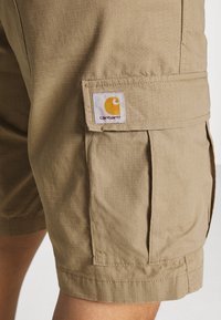 Shorts cargo beiges en tissu léger, avec un motif en grille, des poches latérales et une étiquette logo Carhartt sur la poche avant.