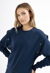 Felpa blu navy con maniche lunghe, scollatura rotonda e bottoni decorativi in tono oro sulle spalle. Tessuto morbido e texturizzato.