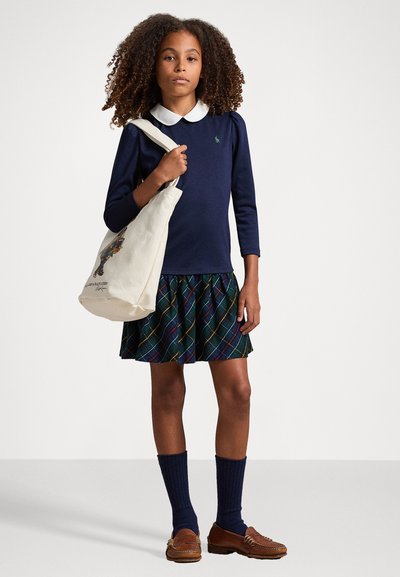 Laivyno spalvos ilgas megztinis su baltu apykakle, raštuota suknele žalios, mėlynos ir raudonos spalvų deriniu, tamsios kelių ilgio kojinės ir rudos odinės loafers tipo batų. Nešioja drobės krepšį.