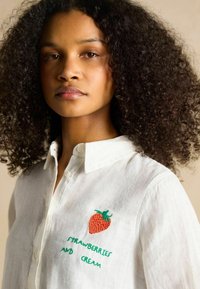 Valge linase särgi peal roheline ja punane maasika tikand ning allpool tekst "strawberries and cream". Klassikaline krae ja nööpidega esiosa.